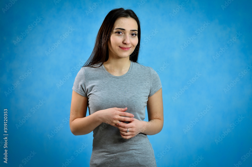 Universal concept of a cute smiling girl on a blue background. Portrait of a pretty young brunette woman in a gray t-shirt. Talking, showing hands with emotions. Universal concept of a cute smiling girl on a blue background. Portrait of a pretty young brunette woman in a gray t-shirt. Talking, showing hands with emotions.