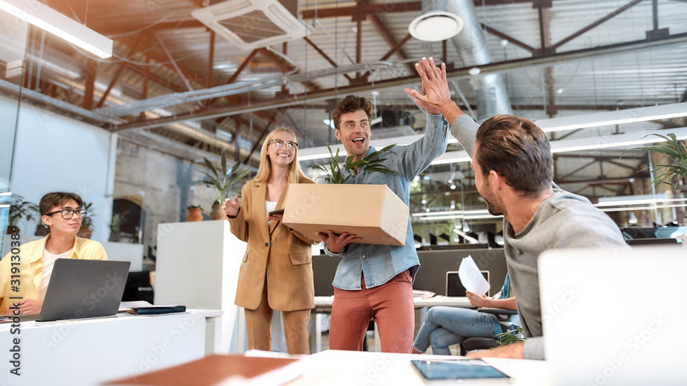 Welcome on board. Young man in casual wear holding box with things and giving high five to his new coworker while standing in the modern office with other colleagues Welcome on board. Young man in casual wear holding box with things and giving high five to his new coworker while standing in the modern office with other colleagues