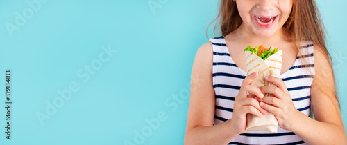 Smiling beautiful child girl eating wrap sandwich. Blue background. Copy space.