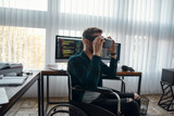 Young male web developer in a wheelchair wearing virtual reality glasses while working at his workplace in modern office. Multiple computer screens with program code