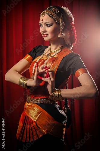 Portrait of white girl as an Indian classical dancer in traditional dress and performing dance performance on the red curtain background. Classical indian temple dance form Bharatanatyam. Dance pose