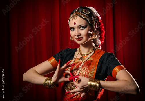 Portrait of white girl as an Indian classical dancer in traditional dress and performing dance performance on the red curtain background. Classical indian temple dance form Bharatanatyam. Dance pose