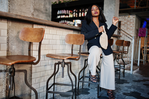 Charming african american woman model in black jacket and waist bag relaxing in cafe during free time.