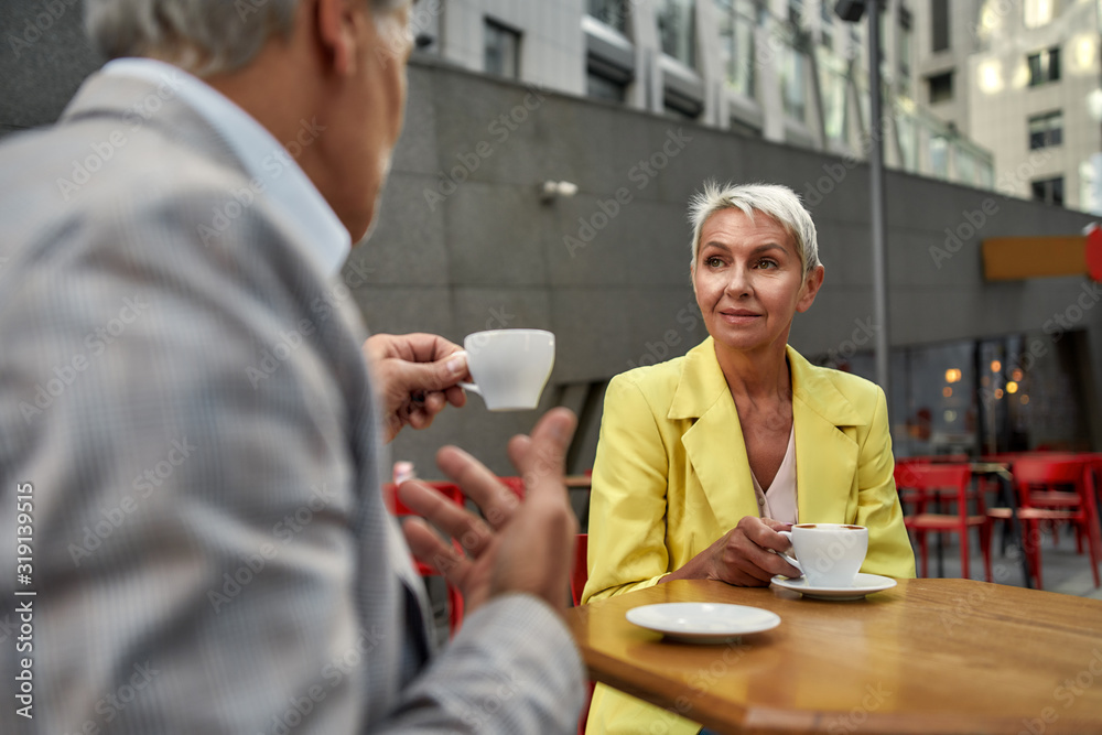 Beautiful middle-aged woman in yellow suit jacket drinking fresh coffee with her friend while sitting in cafe outdoors together