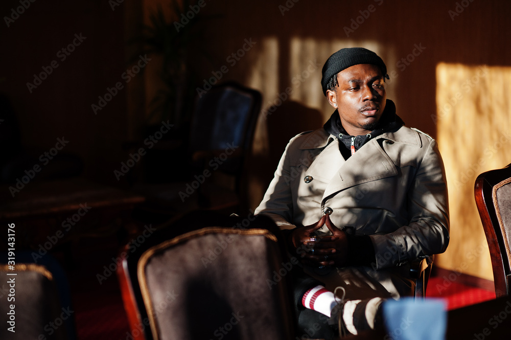 Handsome african american man posing  inside room with sunlight shadows in black hat and beige coat.