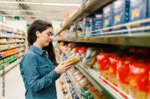 Shopping at the grocery store. A young woman in a denim shirt reads the information on a packet of pasta. In the foreground blurred shelves with products. In profile photo