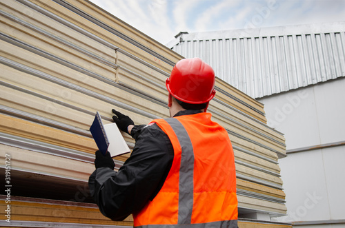  A worker in a protective helmet and overalls makes an inventory of sandwich panels and writes information in his notebook. Pack of Sustainable echo absorber sandwich panel for wall photo