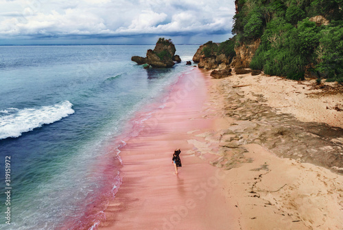 A young woman walks along the sea on a pink beach, Bali, Indonesia. A girl walks along a pink beach in Bali against a stormy sky and rocks, top view.Amazing beautiful pink beach, Bali nature
