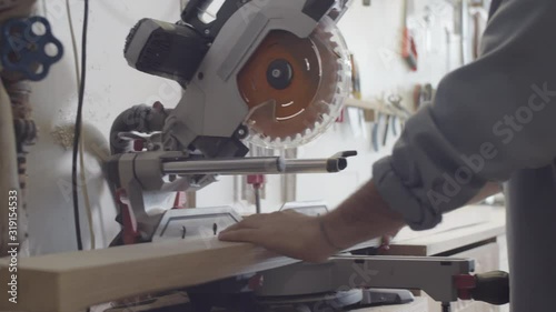 Close-up shot of skilled Caucasian male woodworker carrying wood board to power miter saw, putting it on table, lowering blade and adjusting position against bevels photo