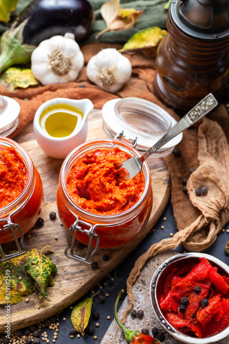 Tabletop scene with raw red chutney with a silver knife surrounded with vegetables. Raw Ajvar in a small glass jar