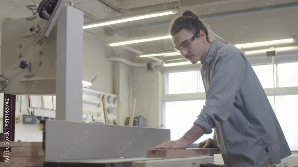Thigh-up shot of young Caucasian carpenter in safety goggles checking surface of wood board then smoothing rough edge on jointer machine, while working at furniture making workshop Thigh-up shot of young Caucasian carpenter in safety goggles checking surface of wood board then smoothing rough edge on jointer machine, while working at furniture making workshop