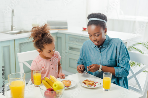african american mother and daughter holding cutlery near tasty pancakes on plates