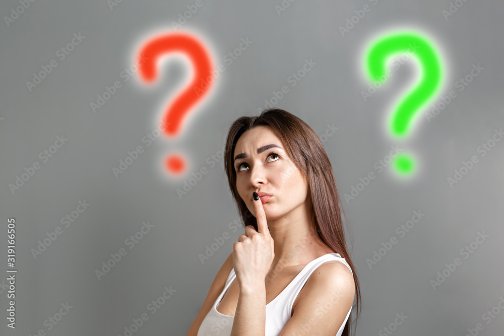 The concept of searching for ideas. Portrait of a pensive young Caucasian woman looking at the question marks at the top and putting her finger to her mouth. Gray background. Copy space The concept of searching for ideas. Portrait of a pensive young Caucasian woman looking at the question marks at the top and putting her finger to her mouth. Gray background. Copy space