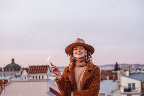 Outdoor winter portrait of elegant happy smiling woman wearing beige hat, turtleneck, brown faux fur coat, holding sparkler, posing in European city. Copy, empty space for text © Victoria Fox