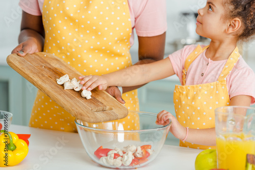 cropped view of african american mother holding cutting board with mushrooms near bowl and cute daughter