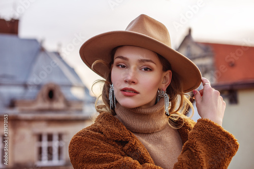 Outdoor close up fashion portrait of elegant woman wearing beige hat, turtleneck, brown coat, trendy big earrings with rhinestones, posing in European city. Copy, empty space for text