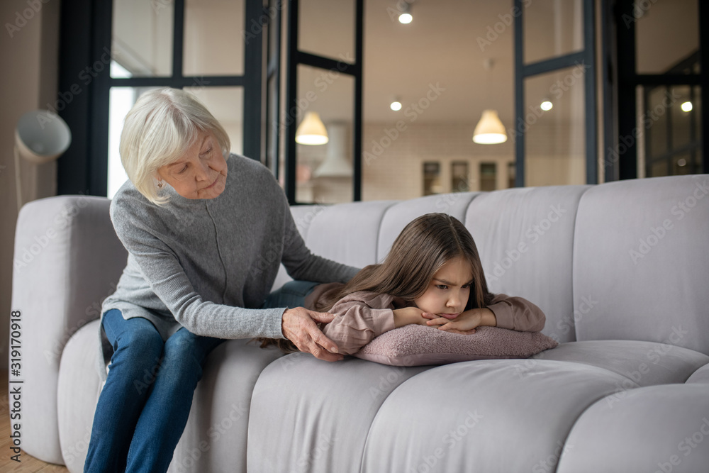 Fototapeta premium Little offended girl lying on the couch and grandmother sitting nearby.
