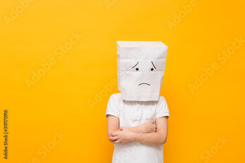 Obraz na płótnie Cute little school boy standing with a paper bag on his head - sad face on yello