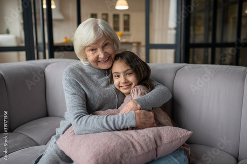 Grandmother and granddaughter hugging and smiling sitting on a sofa.