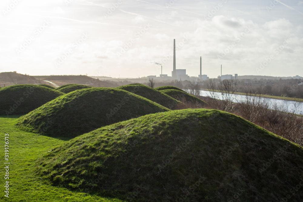 Green small hills in the park. In the background a factory with a large chimney. Denmark