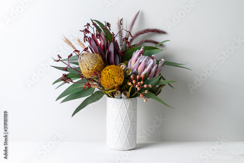 Beautiful floral arrangement of mostly Australian native flowers in a vase, including protea, banksia, kangaroo paw eucalyptus leaves and gum nuts on a white table with a white background. photo