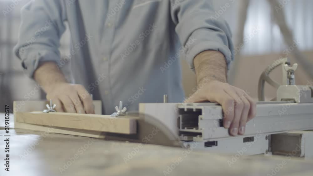 Close-up shot of hands of anonymous carpenter cutting piece of wood fixed to sliding table with clamps, on cabinet saw at woodworking workshop, with sawdust flying around