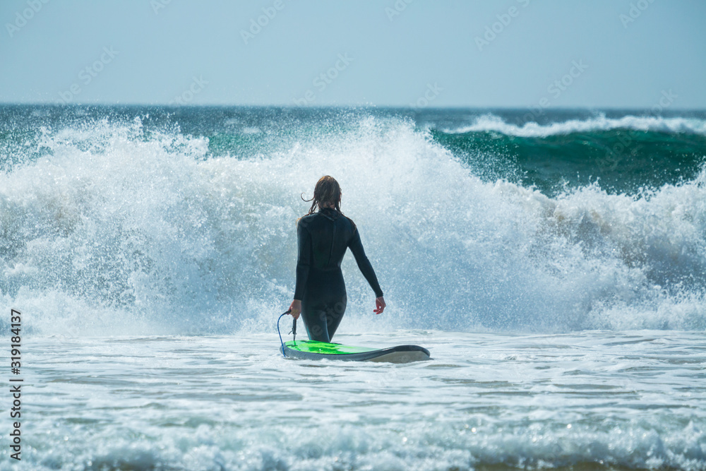 Young woman in black wetsuit holds surfboard and walks into the furious ocean with huge waves Young woman in black wetsuit holds surfboard and walks into the furious ocean with huge waves