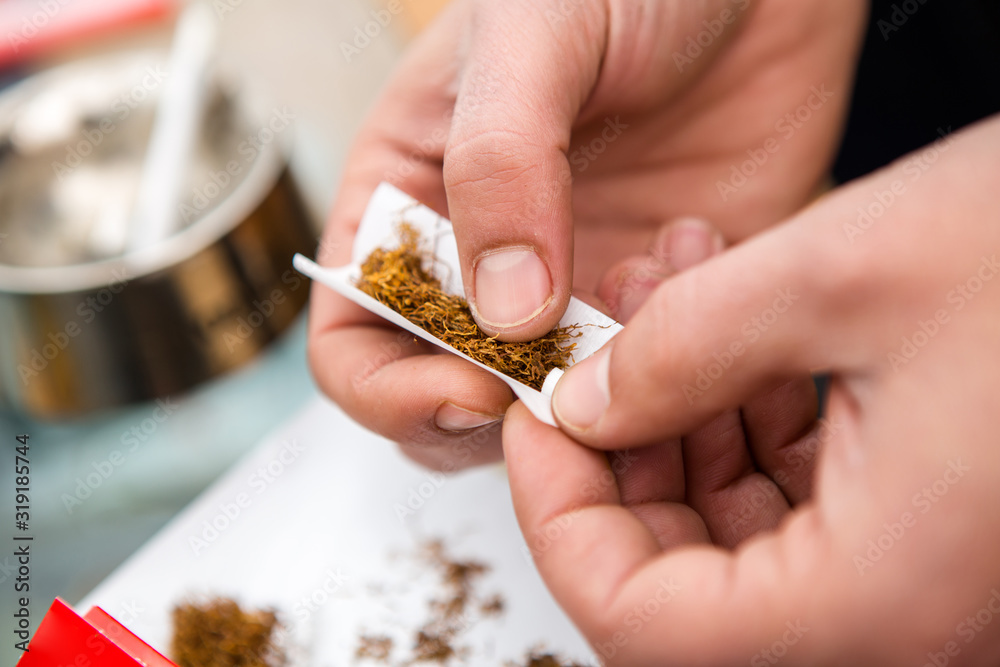 Closeup on the hands of a man just making a tobacco cigarette and a sheet of paper. In the background you can see an ashtray and spilled tobacco. Closeup on the hands of a man just making a tobacco cigarette and a sheet of paper. In the background you can see an ashtray and spilled tobacco.