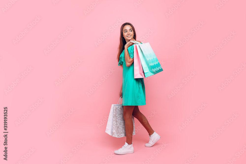 Portrait of a stylish smiling beautiful woman in an off-the-shoulder dress holding bags on an isolated pink background