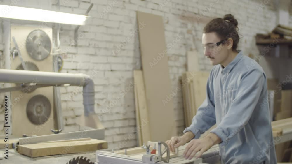 Thigh-up arc shot of young Caucasian carpenter standing in workshop, putting on safety goggles, checking and switching on large cabinet saw and starting to cut piece of wood clamped to sliding table