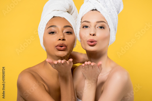 positive multiethnic girls with towels on heads blowing air kisses, isolated on yellow