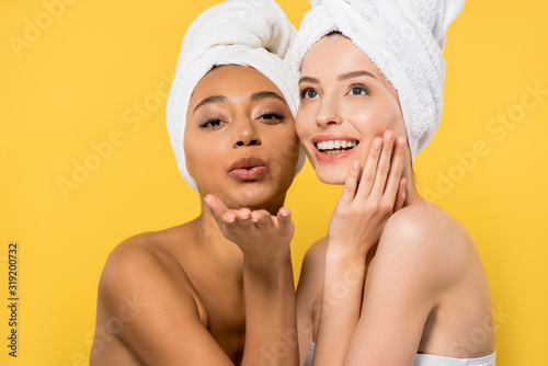 happy multicultural young woman with towels on heads blowing air kiss, isolated on yellow