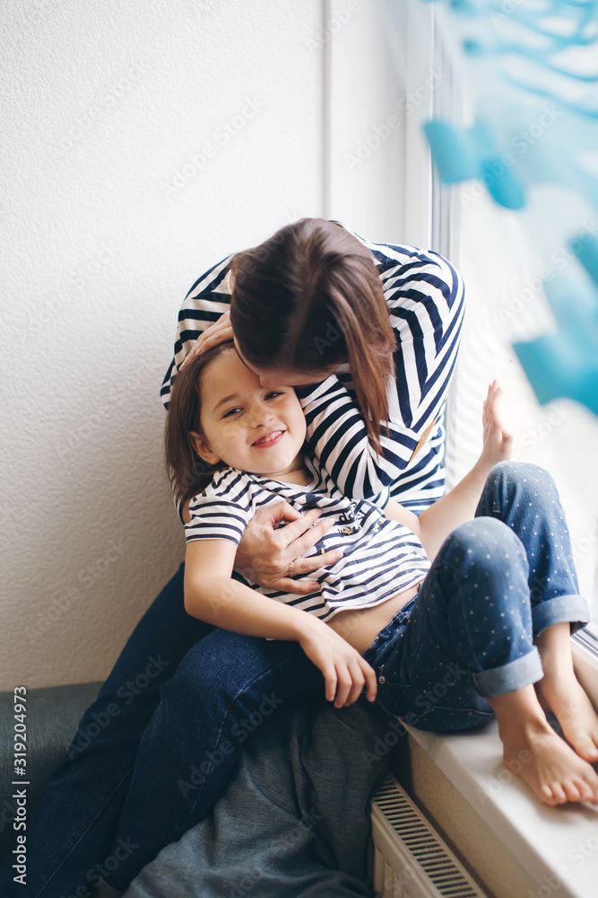 Fototapeta premium Mother and daughter sitting at the window at home hugging and having fun