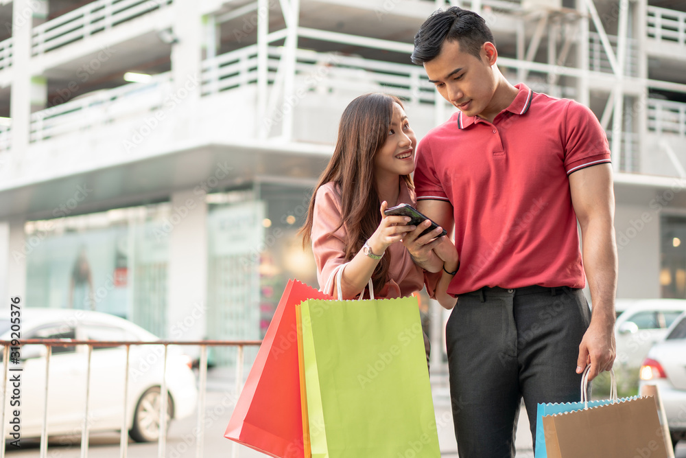 Happy young couple of shoppers walking in the shopping street towards and holding colorful shopping bags in hand and use a smartphone for check promotion. Concept of sale and Black Friday shopping Happy young couple of shoppers walking in the shopping street towards and holding colorful shopping bags in hand and use a smartphone for check promotion. Concept of sale and Black Friday shopping