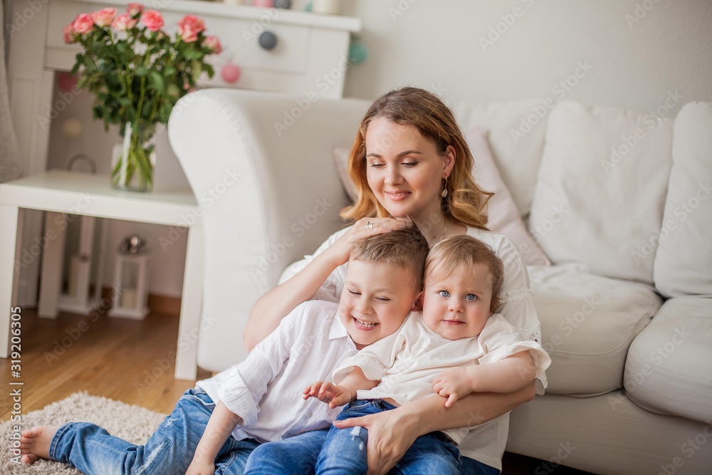 Beautiful young mother, little son, little daughter are sitting on the sofa at home. Mothers Day. A happy family. Cozy.
