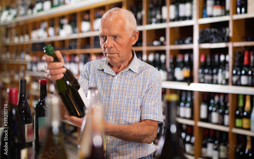 Positive grey-haired elderly man choosing wine in modern wineshop