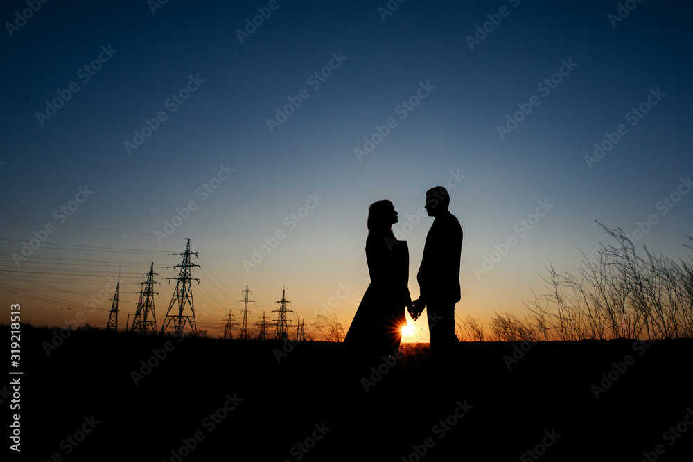Silhouette of Couple hugging on Power lines background on sunset. Save the planet and electricity. Humanity and electricity. Energy Distribution Network - Electricity Pylons. The Eiffel Towers