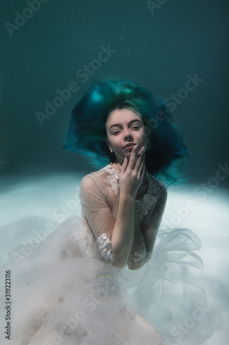 A beautiful girl with blue and long hair swims underwater in the pool in a fluffy white dress. Looks like a nymph or a mermaid