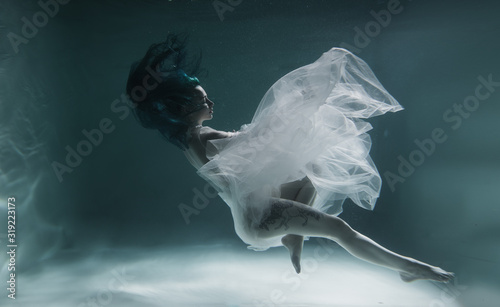 A beautiful girl with blue and long hair swims underwater in the pool in a fluffy white dress. Looks like a nymph or a mermaid