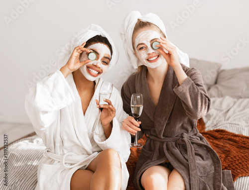 Canvas Print Cheerful girlfriends with face masks wearing bathrobes drinking champagne