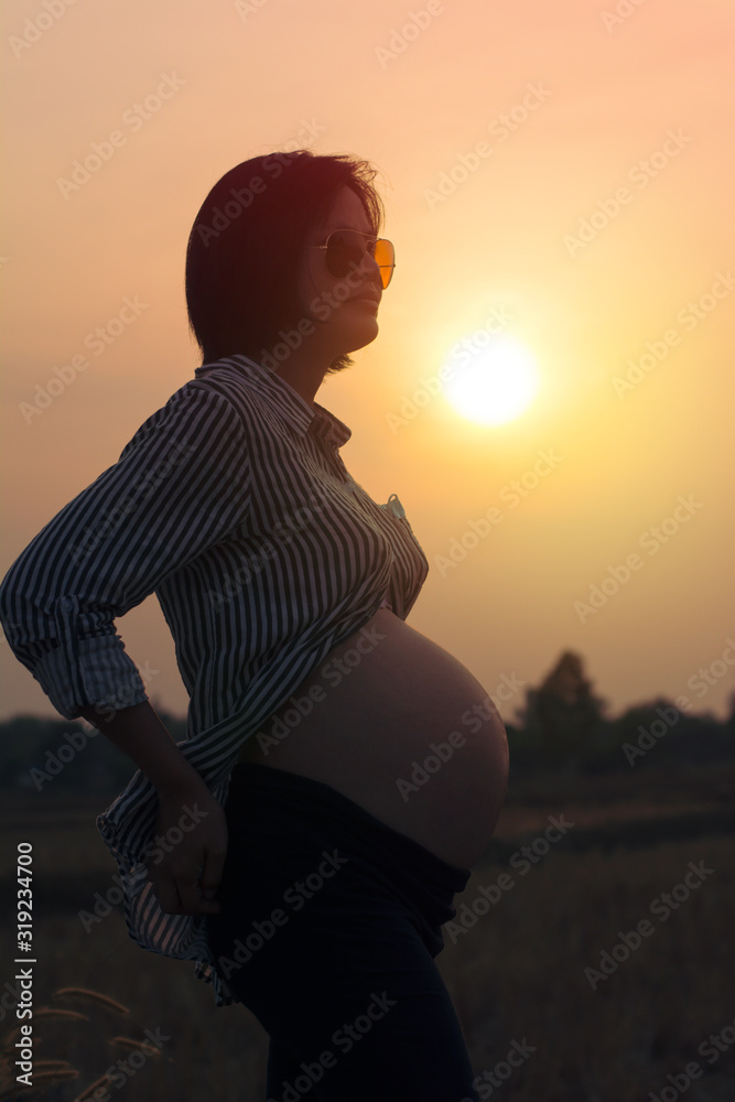 Pregnant women standing on the sunset background. She wears Sunglasses.
