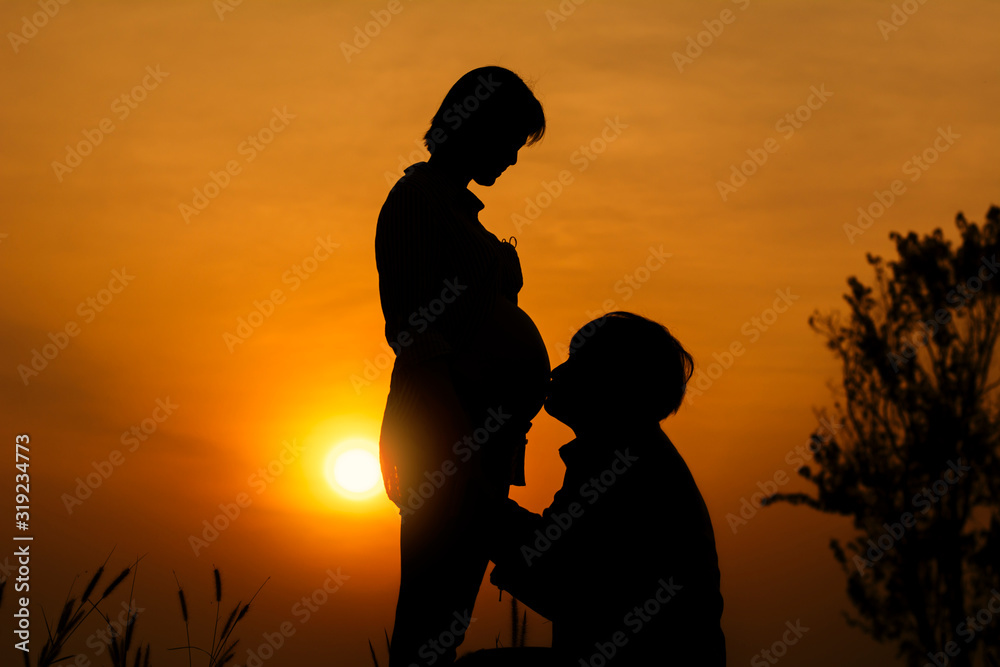 Silhouette of Young man kissing his pregnant wife's belly and talking with their child on sunset background.