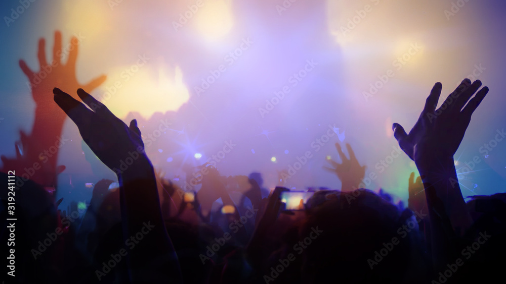 Photo of a concert hall with people silhouettes clapping in front of a big stage lit by spotlights. Shot is taken from concert crowd point of view, lens flare is visible