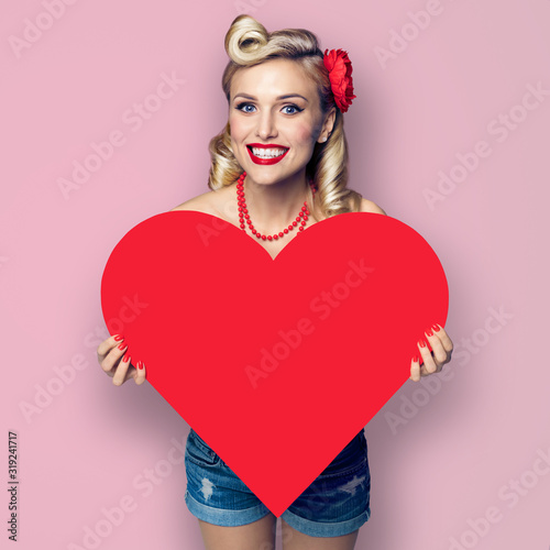 Square composition picture - woman holding red paper heart shape. Pin up girl at retro fashion and vintage concept. Pink background. Copy space for some text. Valentine or Like symbol.