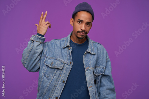 Puzzled young brown-eyed bearded hansome male with dark skin looking confusedly at camera with folded lips, posing over purple background with raised hand
