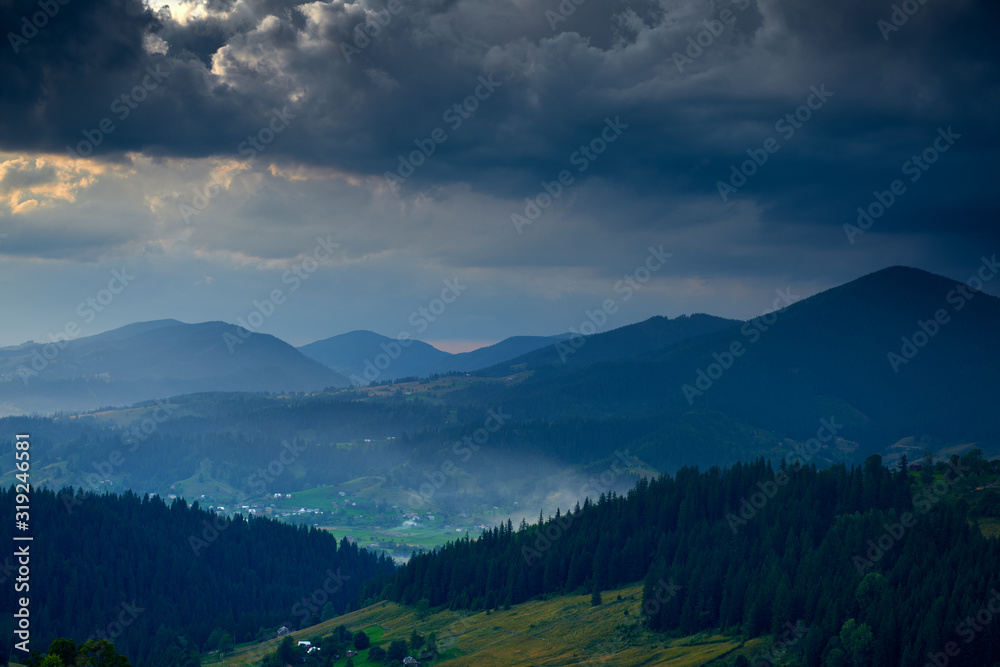 Obraz premium Sunset in carpathian mountains - beautiful summer landscape, spruces on hills, village, homes, dark cloudy sky and bright sun light, meadow and wildflowers