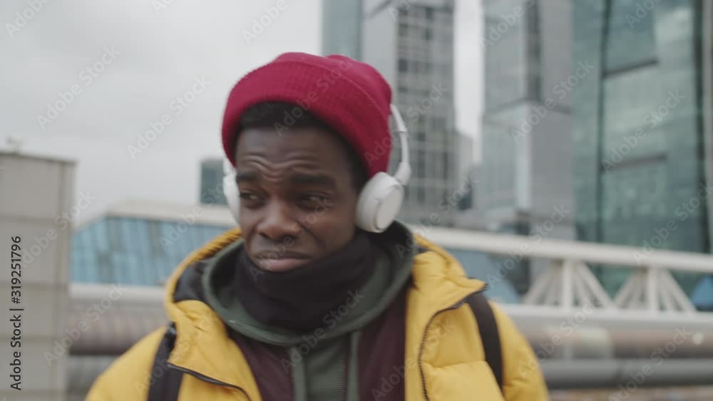 Panning of young African man wearing casual warm clothes walking in city, putting on earphones and starting listening to music Panning of young African man wearing casual warm clothes walking in city, putting on earphones and starting listening to music