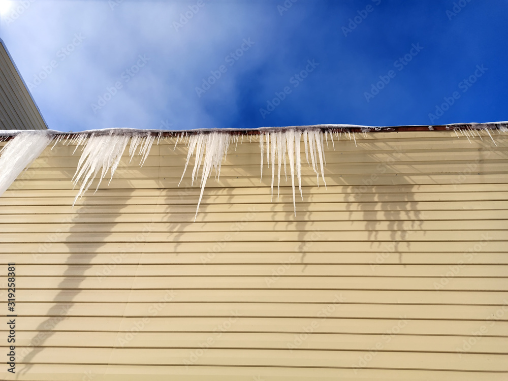 icicles hanging from the roof against a blue sky and a beige wall with shadow and sunbeams icicles hanging from the roof against a blue sky and a beige wall with shadow and sunbeams