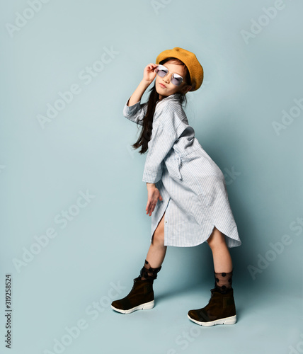 Little asian kid in sunglasses, oversized shirt dress, brown beret, boots. She kissing you, posing on blue background. Full length