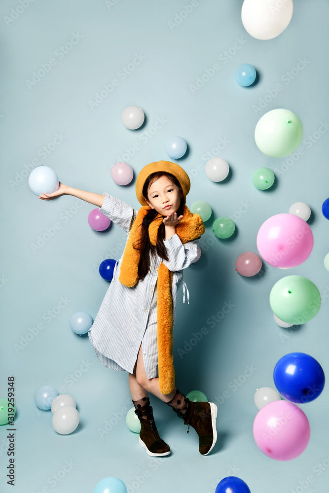 Fototapeta premium Asian child in oversized shirt dress, brown beret and scarf, boots. She sending air kiss, posing with balloons on blue background.